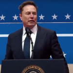 Elon Musk speaking at a public event with a serious expression, wearing a suit and standing behind a podium with the American flag backdrop.