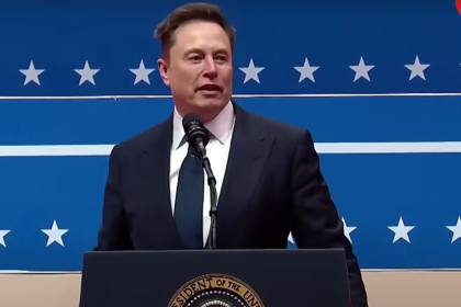 Elon Musk speaking at a public event with a serious expression, wearing a suit and standing behind a podium with the American flag backdrop.