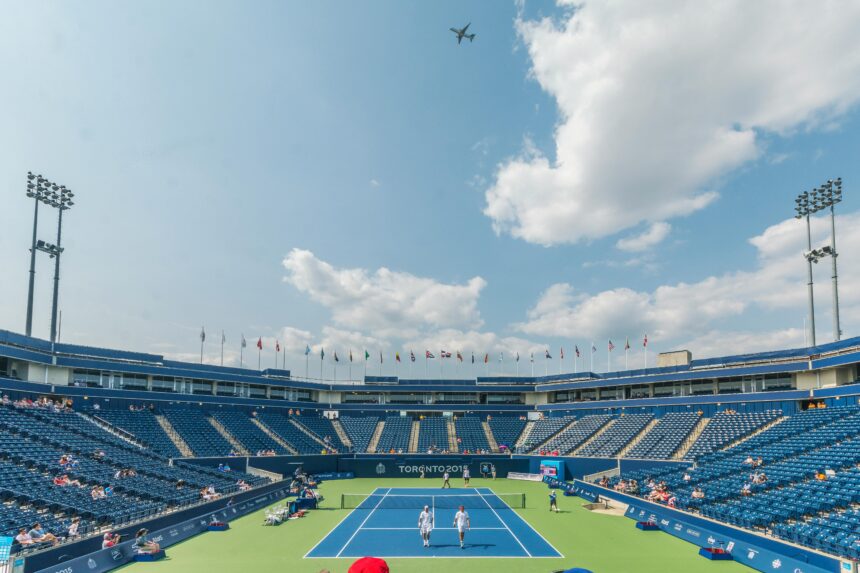 View of a tennis court inside a stadium under a bright blue sky, surrounded by an empty seating area