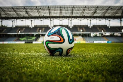 A football on a green pitch with a stadium in the background