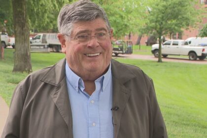 Jym Ganahl smiling outdoors, wearing a brown jacket and light blue shirt, with a green park in the background.