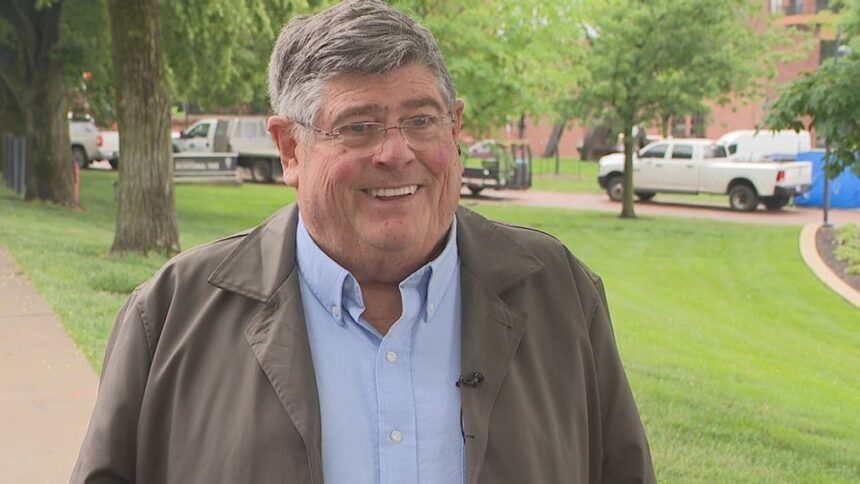 Jym Ganahl smiling outdoors, wearing a brown jacket and light blue shirt, with a green park in the background.