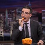 John Oliver on set, holding a food item while delivering a comedic segment on television.