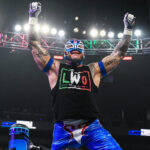 Rey Mysterio raises his arms in victory inside the WWE ring, wearing his signature Lucha Libre mask and LWO (Latino World Order) gear.