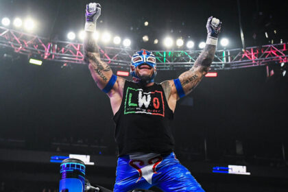 Rey Mysterio raises his arms in victory inside the WWE ring, wearing his signature Lucha Libre mask and LWO (Latino World Order) gear.