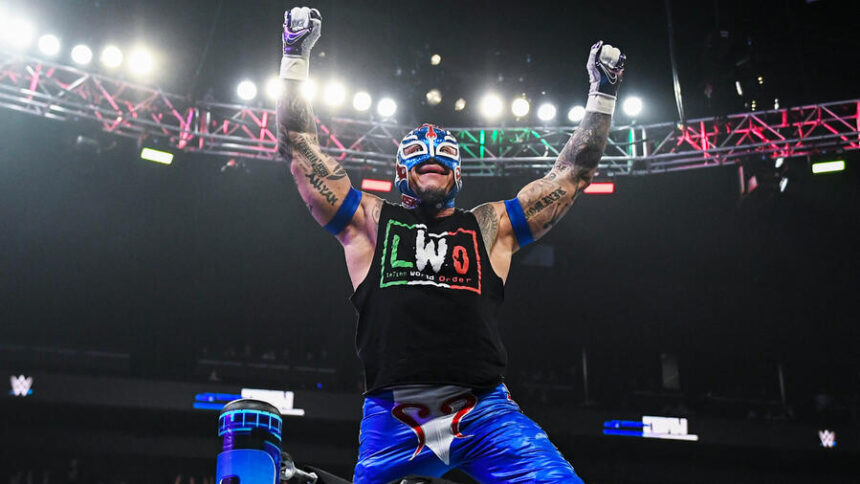 Rey Mysterio raises his arms in victory inside the WWE ring, wearing his signature Lucha Libre mask and LWO (Latino World Order) gear.