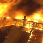 Massive fire engulfs industrial building at night, flames lighting up the sky as smoke billows above the structure.