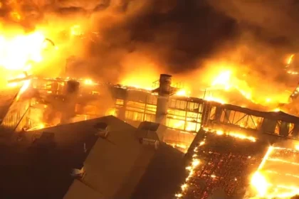 Massive fire engulfs industrial building at night, flames lighting up the sky as smoke billows above the structure.