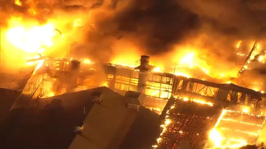 Massive fire engulfs industrial building at night, flames lighting up the sky as smoke billows above the structure.