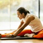 A woman stretching on a yoga mat in a well-lit room, preparing for a workout and recovery session.