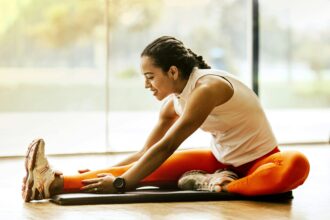 A woman stretching on a yoga mat in a well-lit room, preparing for a workout and recovery session.