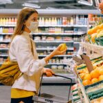 A woman shopping for groceries in a supermarket aisle, selecting fresh produce while pushing a shopping cart.