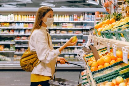 A woman shopping for groceries in a supermarket aisle, selecting fresh produce while pushing a shopping cart.