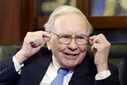 Warren Buffett wearing a suit and tie, smiling as he addresses attendees at the Berkshire Hathaway annual shareholders meeting.